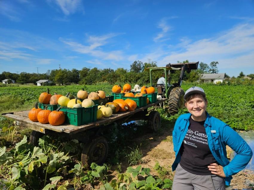 person standing in front of wagon filled with pumpkins in a field